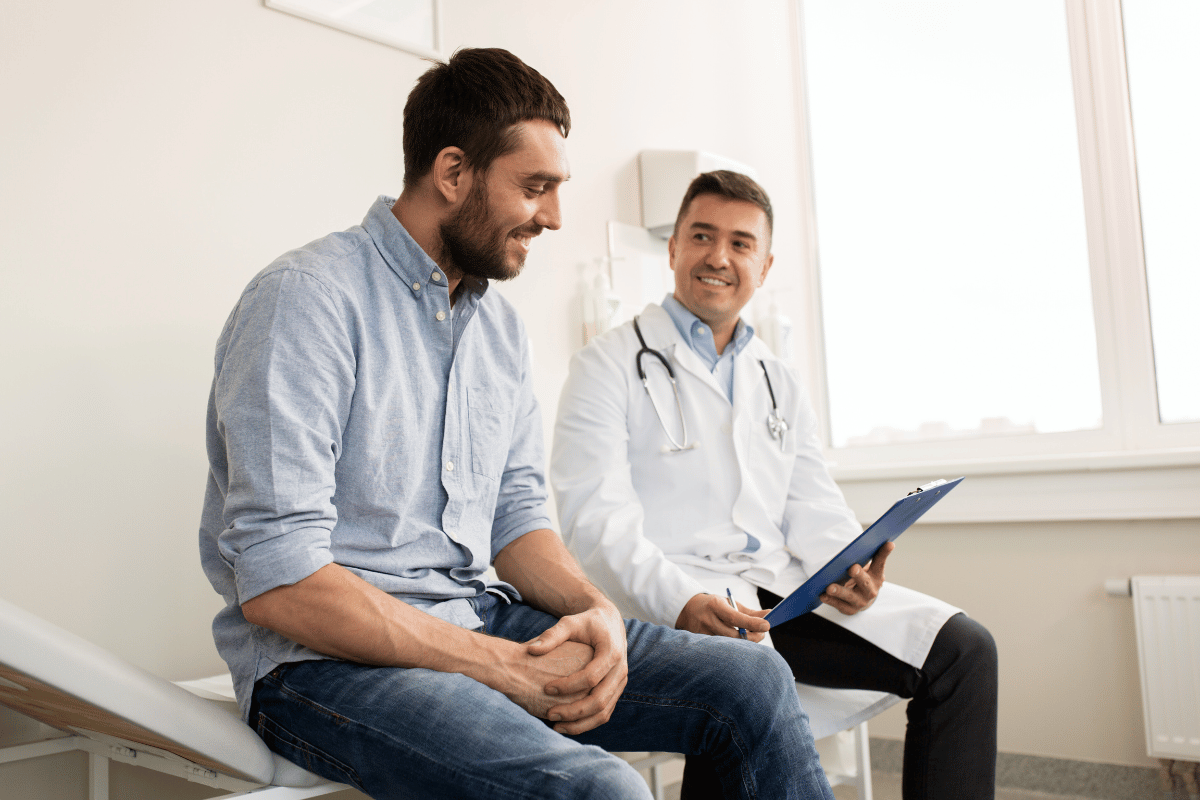 The image shows a doctor consulting with a male patient in a clinical setting. The patient is seated on a medical examination bed, smiling as the doctor discusses something from a clipboard. The atmosphere appears relaxed and friendly, emphasizing trust and communication between the healthcare professional and the patient.