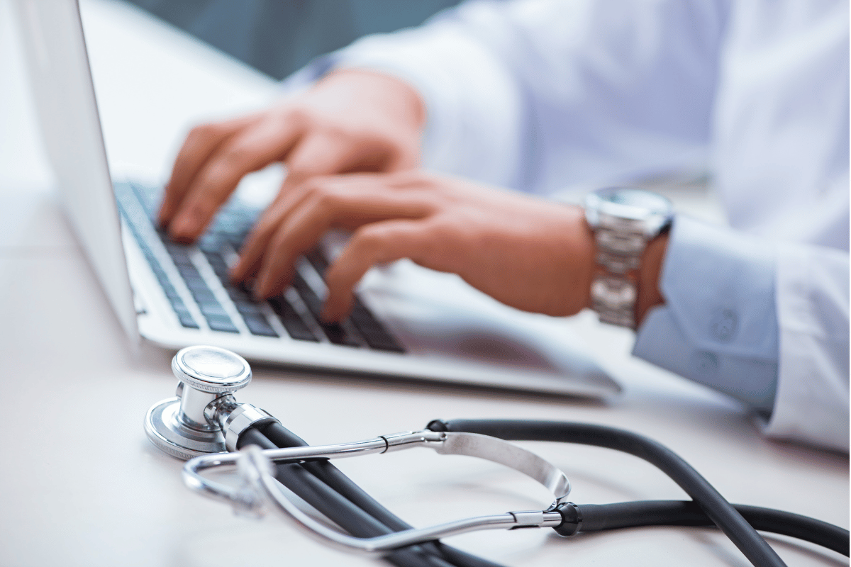 Close-up of a doctor typing on a laptop with a stethoscope placed beside them on the desk.