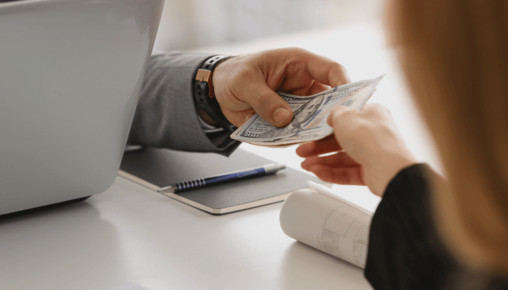 Close-up of a business transaction with hands exchanging US dollar bills, symbolizing getting paid for clinical trials.